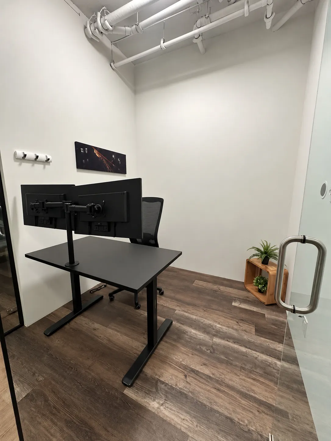 Bureau privé 104-2 au 2727 Coworking, Griffintown : bureau noir avec bras double écran, fauteuil en filet, œuvre d'art et plante dans une caisse de bois