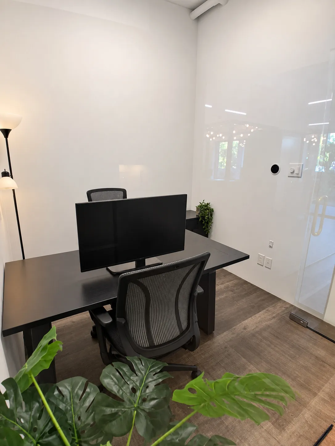 Bureau privé 104-3 au 2727 Coworking, Griffintown : bureau noir avec écran et deux chaises en maille, lampe sur pied et monstera, près de la façade vitrée.