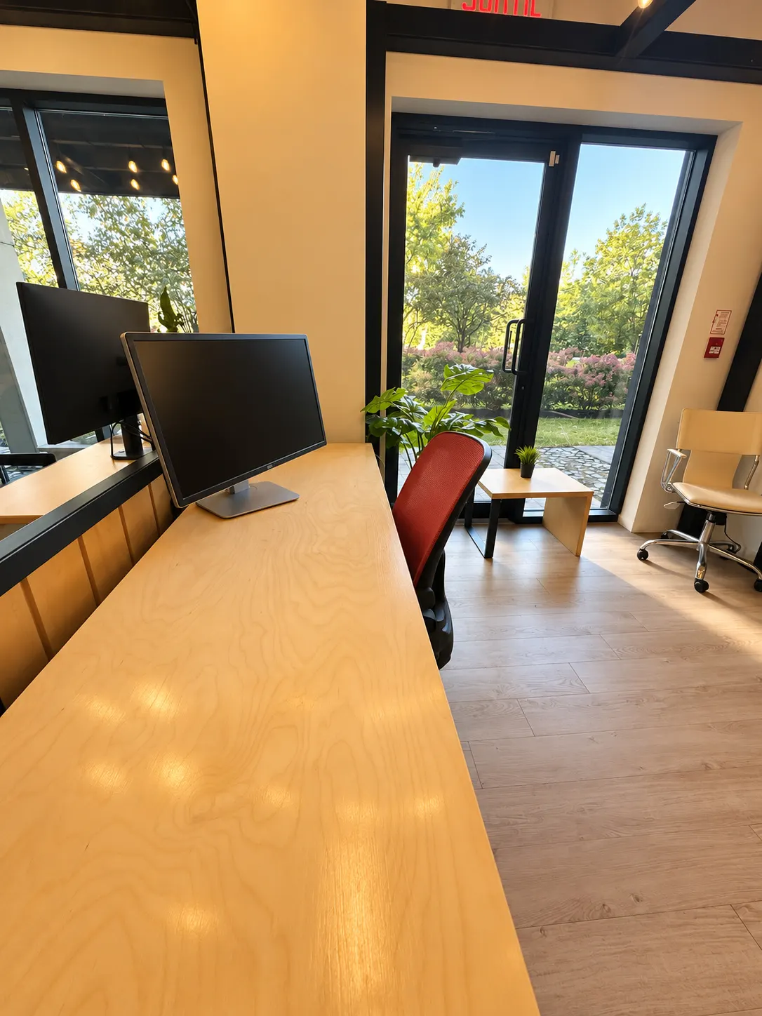 Poste dédié 105-EF au 2727 Coworking, Griffintown : long comptoir en bouleau et chaise rouge, à côté d'une porte vitrée qui donne sur les arbres et les arbustes en fleurs